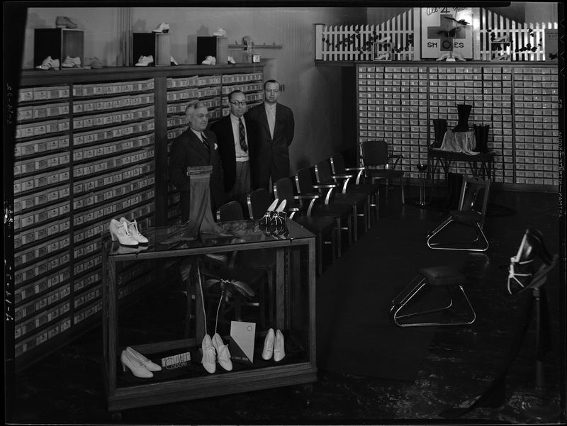 Three unidentified men are standing inside of Murphy Clo Company Bootery. There are shoes on display and shoe boxes line the shelves on the walls. There is a small area with chairs in the middle of the room for people to try on shoes.