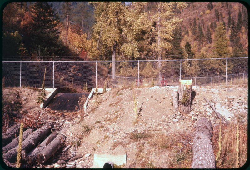 Color photo of a small body of water, probably a canal. There are trees in the area. The water source is fenced in.