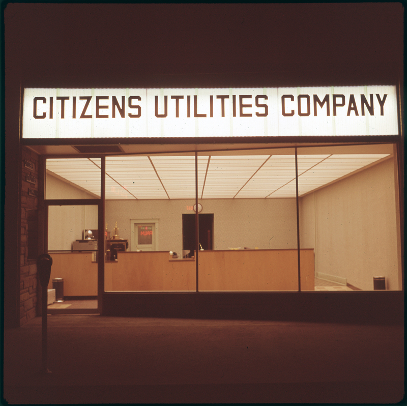 Photo of the exterior of the Citizens Utilities Company building. The lights are on inside of the building and there is a small window display and a Christmas tree. There is also a car parked in front of the building that is visible.