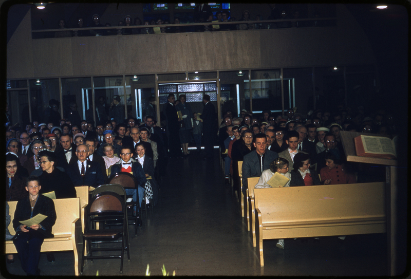 Color photo of the Congregational Church dedication gathering. Men, women. and children are sitting in the pews facing away from the photographer. There are people seated on stage as well, possibly the choir.