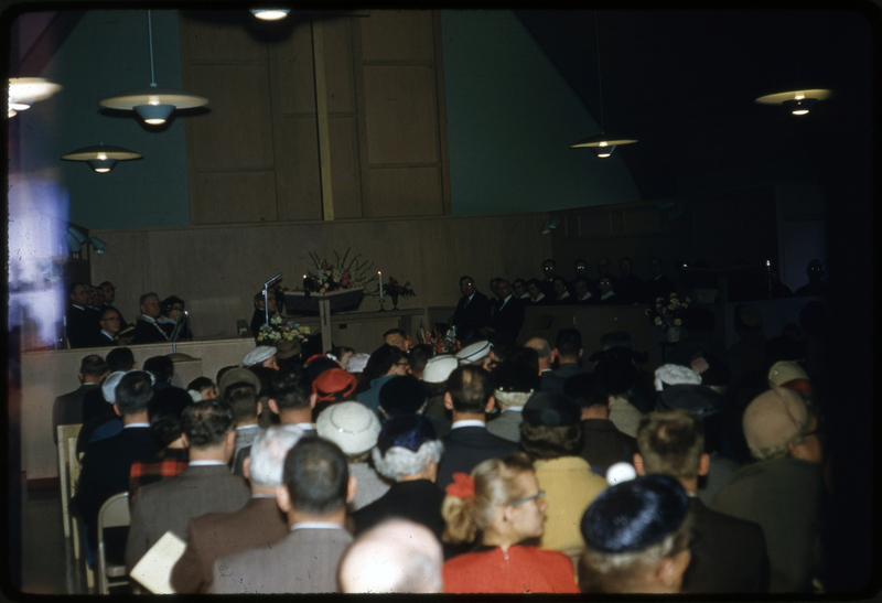 Color photo of the Congregational Church dedication gathering. Men, women. and children are sitting in the pews and looking at the photographer.