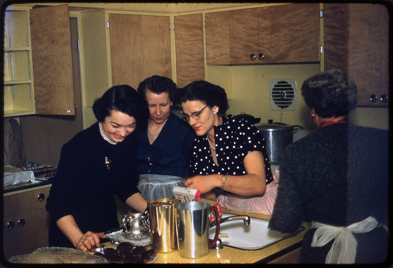 Color photo of four women in the kitchen, three of which are gathered around the counter to look at the food.