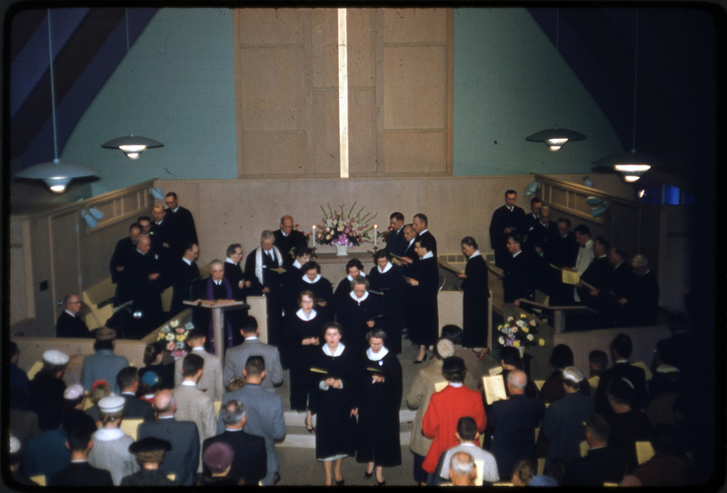 Color photo of people walking off the stage of the Congregational church dedication gathering. They are likely the choir. They are walking off the stage and past the crowd in the pews.