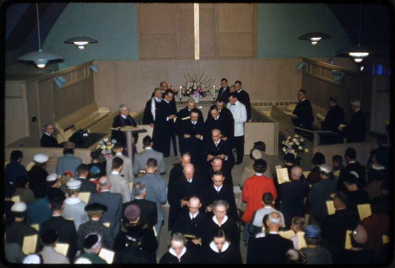 Color photo of people walking off the stage of the Congregational church dedication gathering. They are likely the choir. They are walking off the stage and past the crowd in the pews.