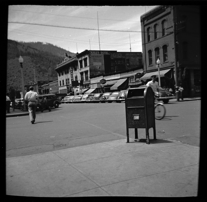 Photo of downtown Wallace, Idaho. There is a man riding a bicycle on the street and another walking away from the photographer. Several buildings are visible and cars are parked on both sides of the road.