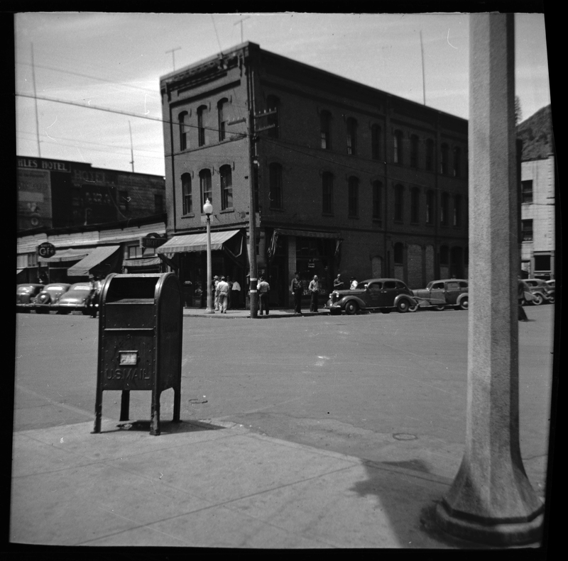 Photo of downtown Wallace, Idaho. A group of unidentified men stand on the street corner opposite of the photographer outside of a building. There are cars parked on the side of the road and a mailbox in front of the photographer.