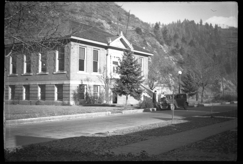 Photo of the Wallace Carnegie Public Library. There is a car parked out front and two people standing next to it. The building appears to be two stories and has several trees planted around it.