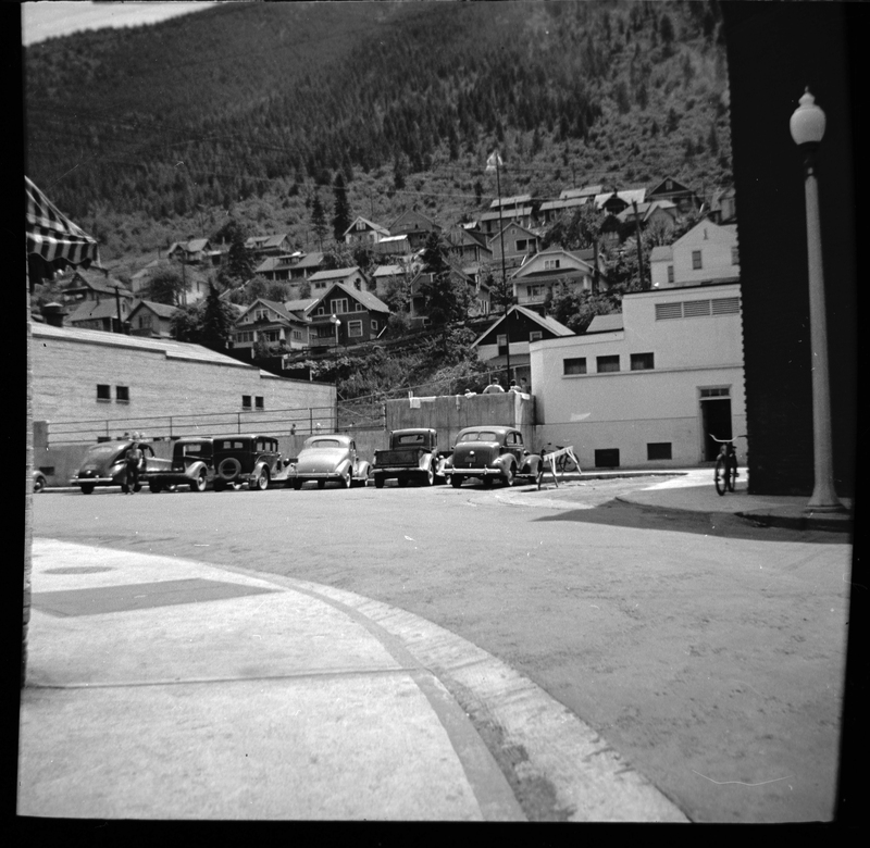 Photo of a street scene in Wallace, Idaho. There is a building with cars parked in front of it directly in front of the photographer, and houses can be seen further up a hill behind that building.