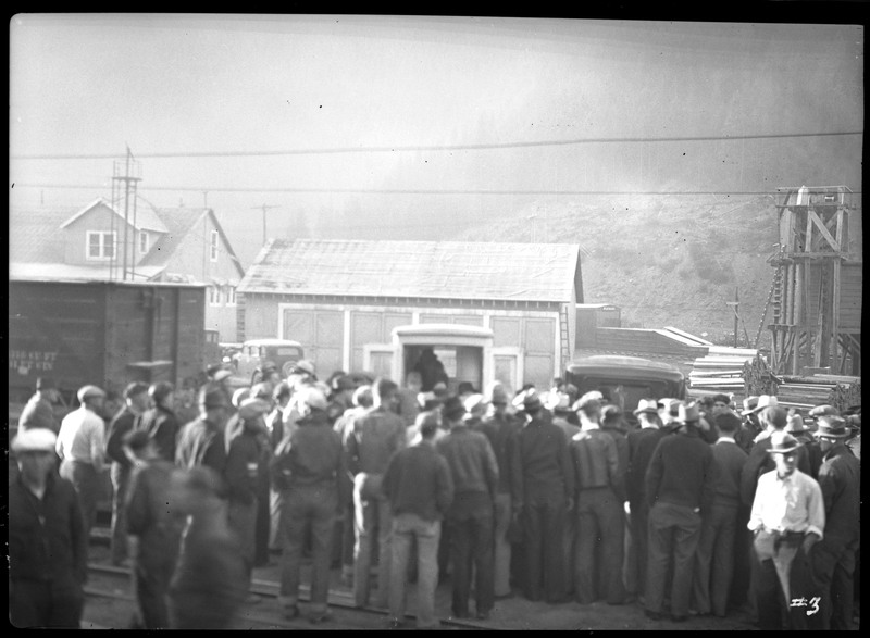 People gathered on railroad tracks in front of Morning Mine buildings in Mullan, Idaho. There are cars in the background. 