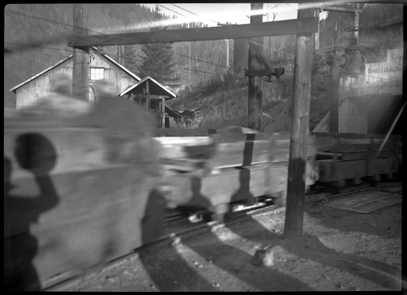 Mine cars emerging from the mine entrance. There are shadows of onlookers visible. What is visible of the inscription above the mine entrance reads "19", "MORNIN", "FEDERAL".
