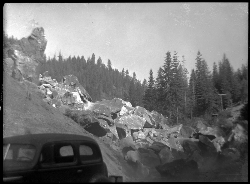 The aftermath of the blast. The dust has settled, and large chunks of rock are visible, piling at the bottom of the hill. There is a car in the foreground. 