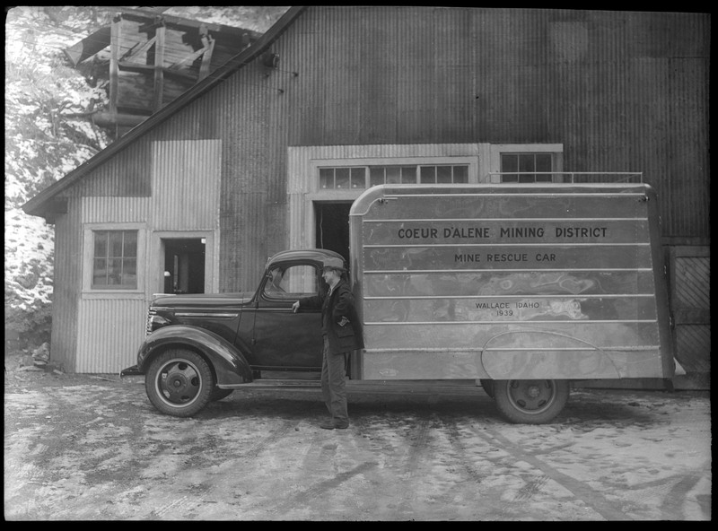 An unidentified man posing in front of a mine rescue vehicle. The side of the vehicle reads, "Coeur d'Alene Mining District; Mine Rescue Car; Wallace, Idaho; 1939." There is a building in the background.