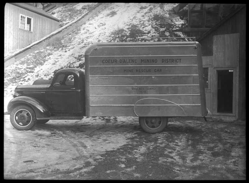 Mine rescue vehicle.  The side of the vehicle reads, "Coeur d'Alene Mining District; Mine Rescue Car; Wallace, Idaho; 1939." There are buildings in the background.