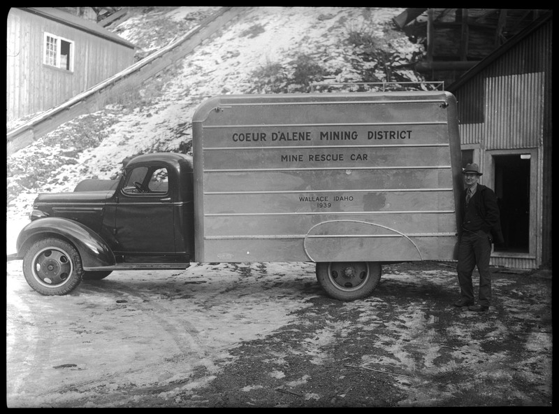 A man posing next to a mine rescue vehicle.  The side of the vehicle reads, "Coeur d'Alene Mining District; Mine Rescue Car; Wallace, Idaho; 1939." There are buildings in the background.