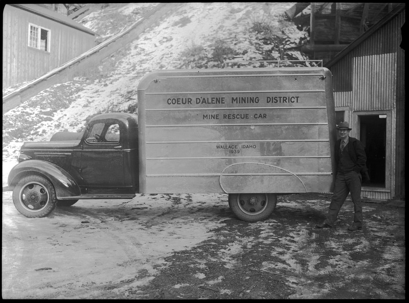 A man posing next to a mine rescue vehicle.  The side of the vehicle reads, "Coeur d'Alene Mining District; Mine Rescue Car; Wallace, Idaho; 1939." There are buildings in the background.