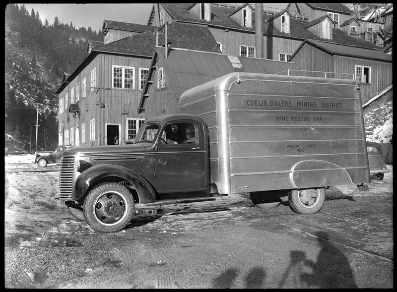 A mine rescue vehicle. The side of the vehicle reads, "Coeur d'Alene Mining District; Mine Rescue Car; Wallace, Idaho; 1939." There are buildings and cars in the background. In the foreground, the shadow of the person taking the picture is visible.
