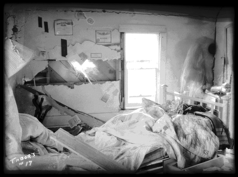 Damage done to a bedroom in N. J. Osborne's house by the Sunshine Mine powderhouse explosion. The walls are broken and cracked and there is debris all over the room and bed.