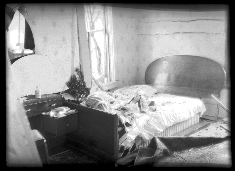 Damage done to a bedroom in N. J. Osborne's house by the Sunshine Mine powderhouse explosion. The walls and window are damaged, and there is debris all over the room and bed.