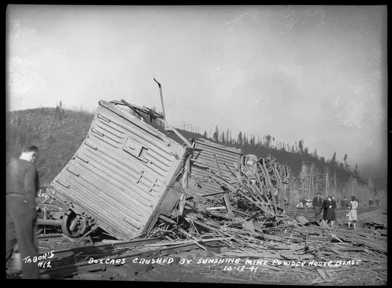 Boxcars crushed by the Sunshine Mine powderhouse explosion. Debris is spilled over the railroad tracks and the surrounding area from the boxcar. Several people are standing in the area inspecting the damage.
