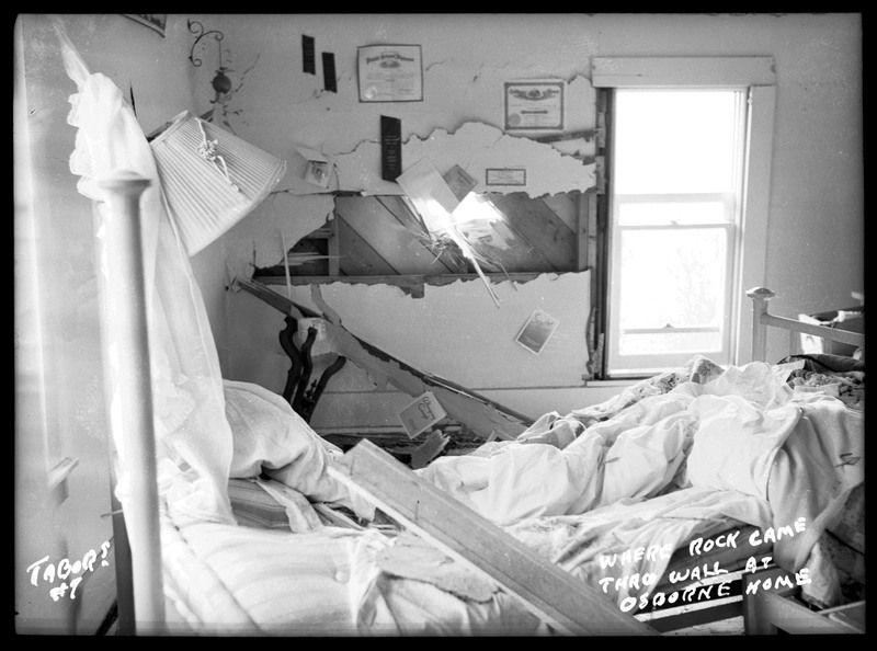 Image depicting where a large rock was thrown through a wall of the Osborne home by the Sunshine Mine powderhouse explosion. The room is extremely damaged and there is a bed with various debris in the room.