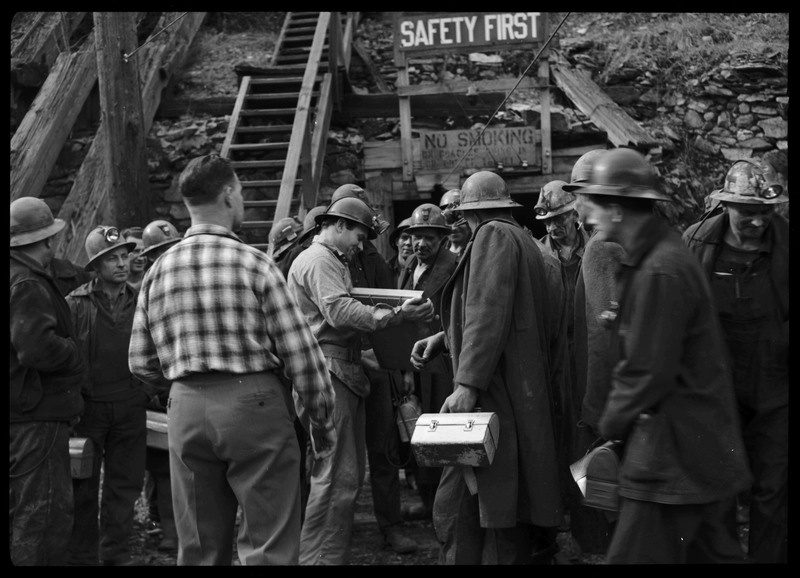 Group of miners from Hecla Mining Company showing off their recently granted safety award to each other. There are signs at the mine entrance behind everyone that read, "SAFETY FIRST" and "NO SMOKING ON COACHES GOING IN OR OUT OF TUNNEL."