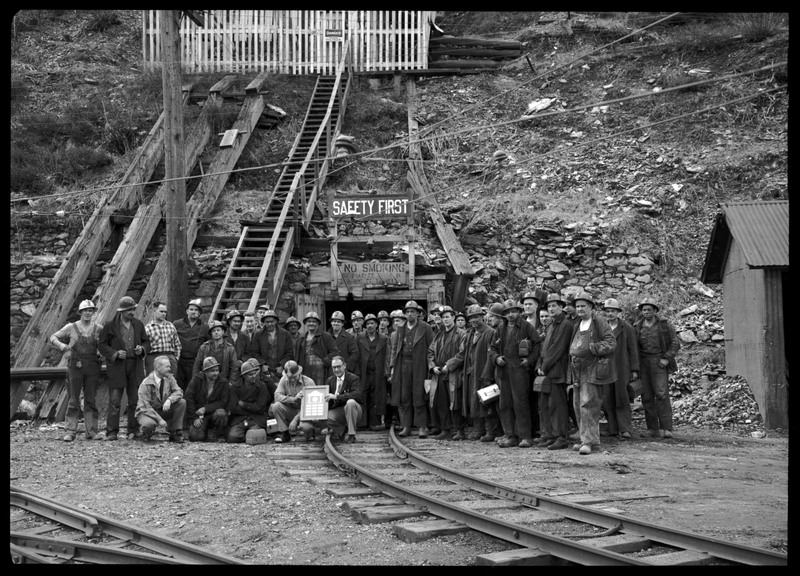 Hecla Mining Company miners posing together for a photo with their recently granted safety award. They are all standing outside of the mine entrance that has two signs posted. One reads, "Safety First," and the other reads, "No Smoking on coaches going in or out of tunnel."