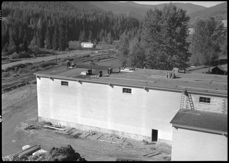 The exterior of Lucky Friday mill facilities and buildings which are under construction. Workers are on the roof of a building. A billboard on the side of the road reads, "WELCOME TO IDAHO".