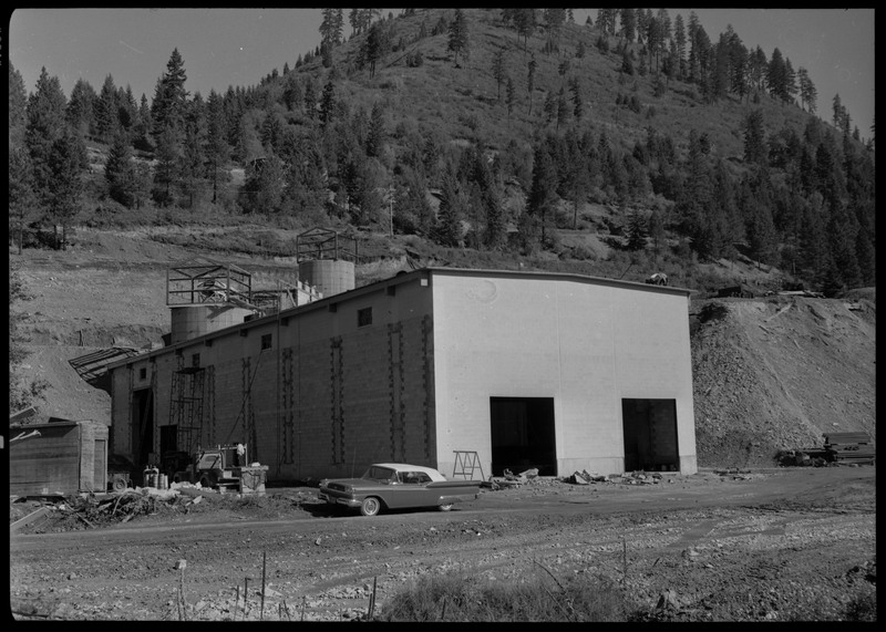 The exterior of Lucky Friday mill facilities and buildings which are under construction, including what appears to be silos for holding mineral materials. Workers are on the roof of a building. A car is parked in front of a building. 