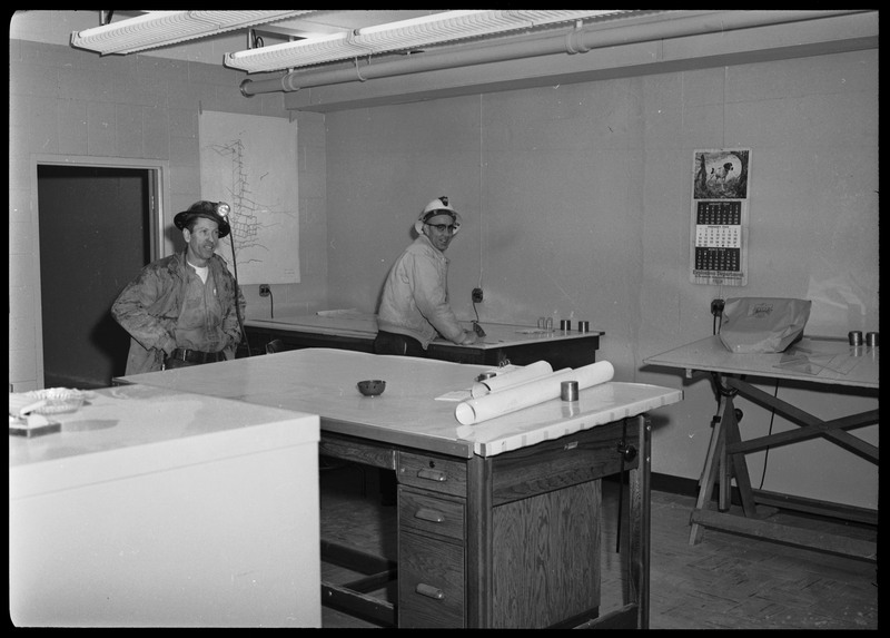 Two miners wearing hard-hats standing in a room with multiple desks. What appears to be a map of the Lucky Friday mine shaft hangs on a wall behind them. At the bottom of a calendar hanging on a wall is an inscription that reads "Explosives Department".