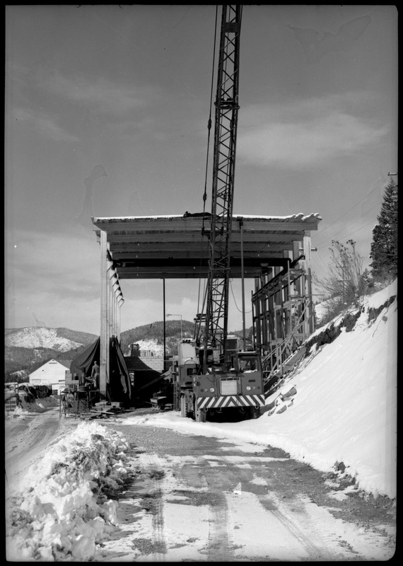 A large crane that is outside of the Lucky Friday Mine buildings. There is snow on the ground surrounding the crane and the small shelter that is partially over it, which has a roof but no walls.