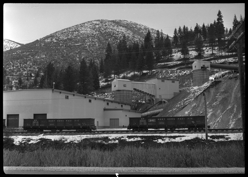 The exterior of the Lucky Friday mine exterior. There are two unattached Northern Pacific train cars on the railroad tracks between the building and the photographer. There is also snow on the ground.
