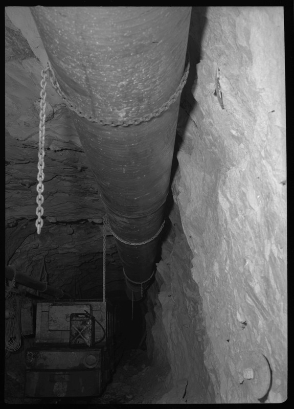 Image of a long pipe that is hung up underground at Bunker Hill. The pipe is held up by chains attached to the rocky ceiling and walls.