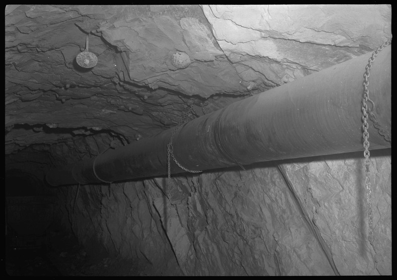 Image of a long pipe that is hung up underground at Bunker Hill. The pipe is held up by chains attached to the rocky ceiling and walls.