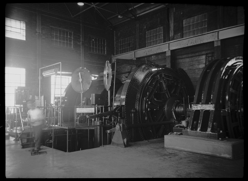 Image of the Hecla Mine mine hoist. There is a blurry figure of a person walking by the machinery. There is a sign on the hoist deck that reads, "Talking to hoistman while hoist is in motion is forbidden; No admission to hoist deck."
