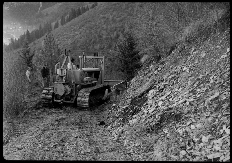 Group of men operating a piece of machinery in a heavily wooded area. They are all standing near the machine and appear to be planning on how to move it.