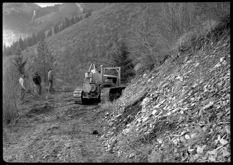 Group of men operating a piece of machinery in a heavily wooded area. They are all standing near the machine and appear to be planning on how to move it.