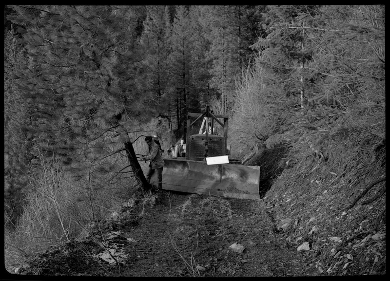 A couple men moving a piece of machinery through a heavily wooded area. They appear to be stuck due to a tree stump that is on the path.