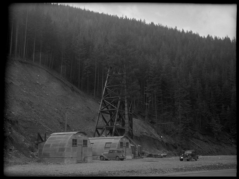 Image of the Rock Creek Mine tower. Notes say that Rock Creek Mine is 4 miles east of Wallace, Idaho. There are two cars parked nearby the tower and buildings.