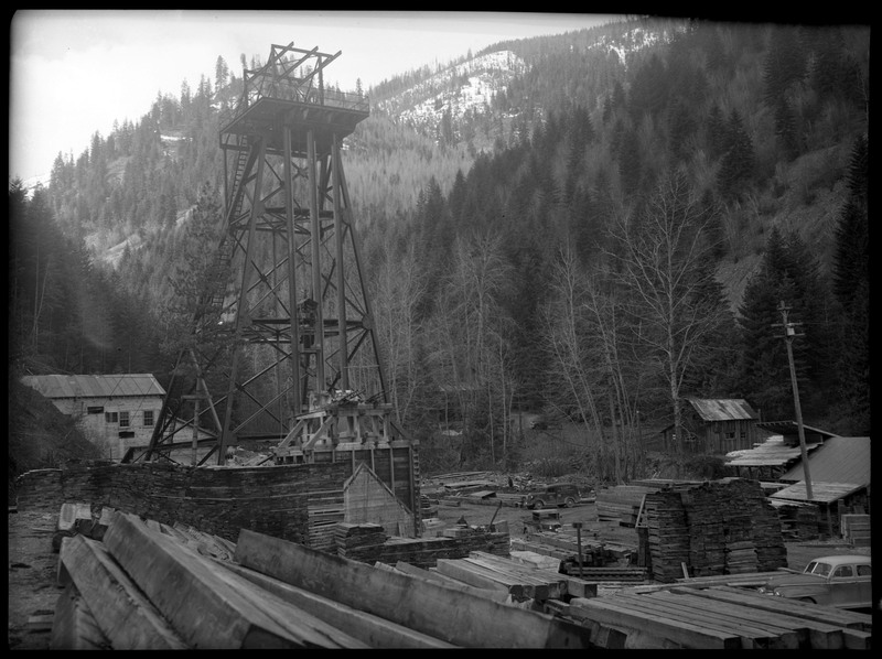Image overlooking Rock Creek Mine. Notes say that Rock Creek Mine is located 4 miles east of Wallace, Idaho. The tower, several buildings, construction supplies, and cars are all visible in the small area surrounded by trees.