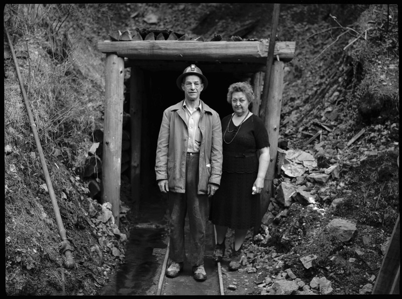 An unidentified man and woman standing outside in front of the entrance to Silverton Mine in Silverton, Idaho. The man is wearing a hard hat, suggesting he is a miner.
