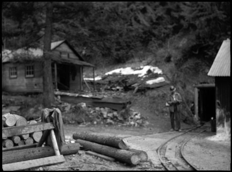 An unidentified man walking out of the entrance to Silverton Mine. The photo is slightly out of focus, with the man and the building to the left blurry. There is some chopped wood on the ground that is in focus.