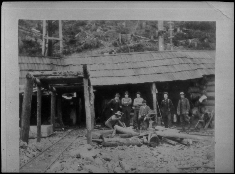 Image of a photograph of a group of men gathered together at Hercules Mine in Burke, Idaho. The men are all standing outside of a building which is likely the mine entrance. One of the men appears to be hitting something with a pickaxe.