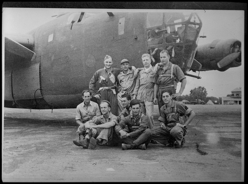 Image of a photograph of nine men posing for a photo in front of a large military aircraft. The aircraft is likely a B-24 Liberator.