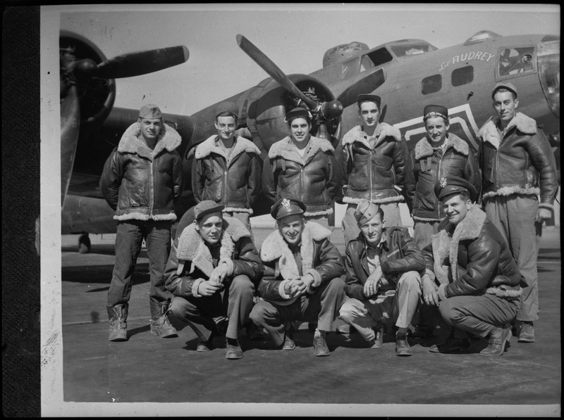 Image of a photograph of ten men posing in front of a military aircraft. All of the men are wearing matching jackets and similar hats. The aircraft is a B-17 named Lil' Audrey.