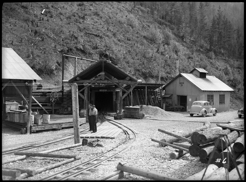 Two men standing outside the mine entrance at Silver Summit. There is a car parked next to one of the buildings, and railroad tracks for the mine carts.