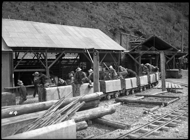 A group of miners getting out of the mine carts at the entrance of Silver Summit mine. There is a building behind them and the entrance of the mine is visible.