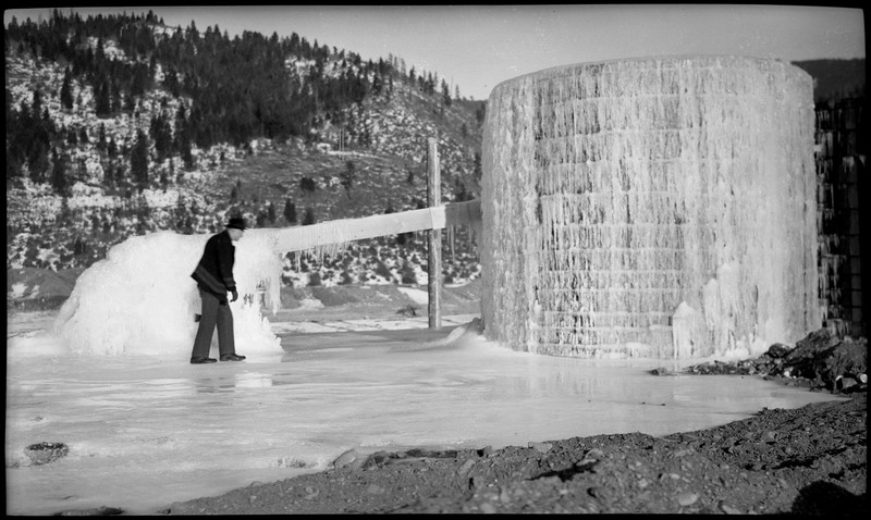 An unidentified man walking on a large patch of ice near a mill that is completely frozen over in ice.