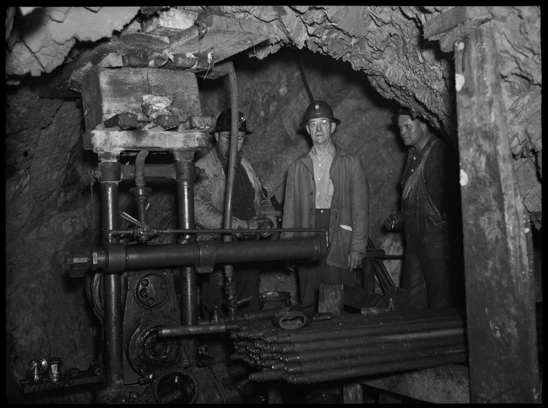 Three men working inside of an unidentified mine. They are all standing in a tunnel behind a large piece of machinery.