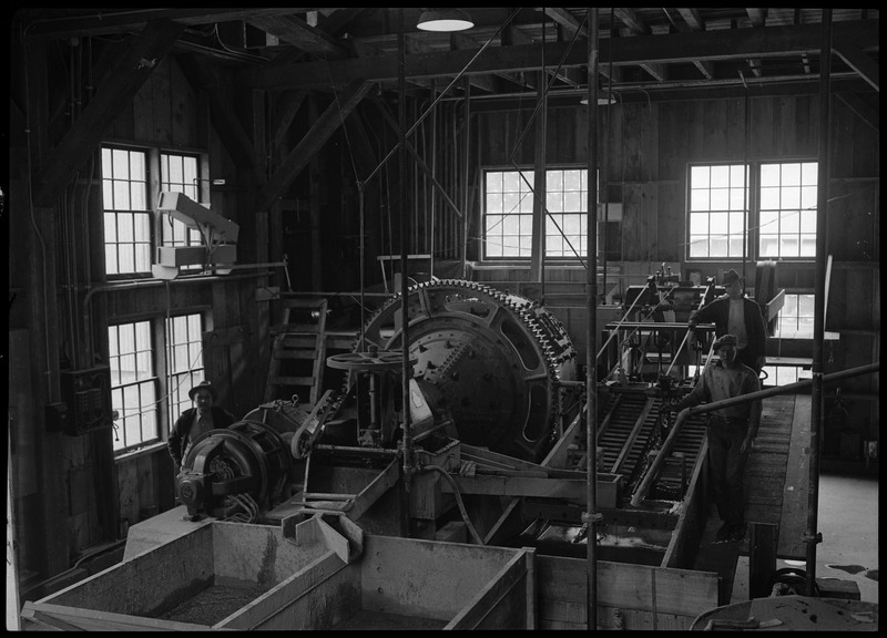 Two men standing next to milling equipment inside of an unidentified milling complex.