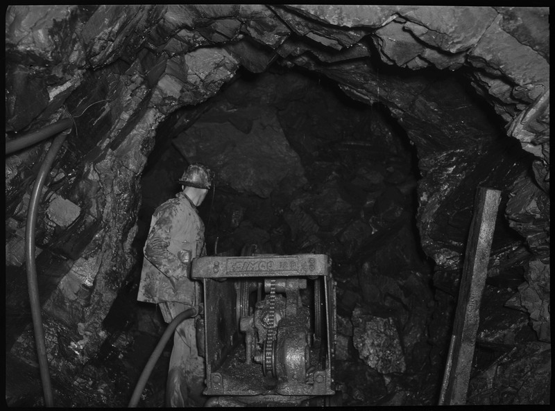 A man working inside of an unidentified mine. He is standing in a tunnel and operating a piece of machinery.